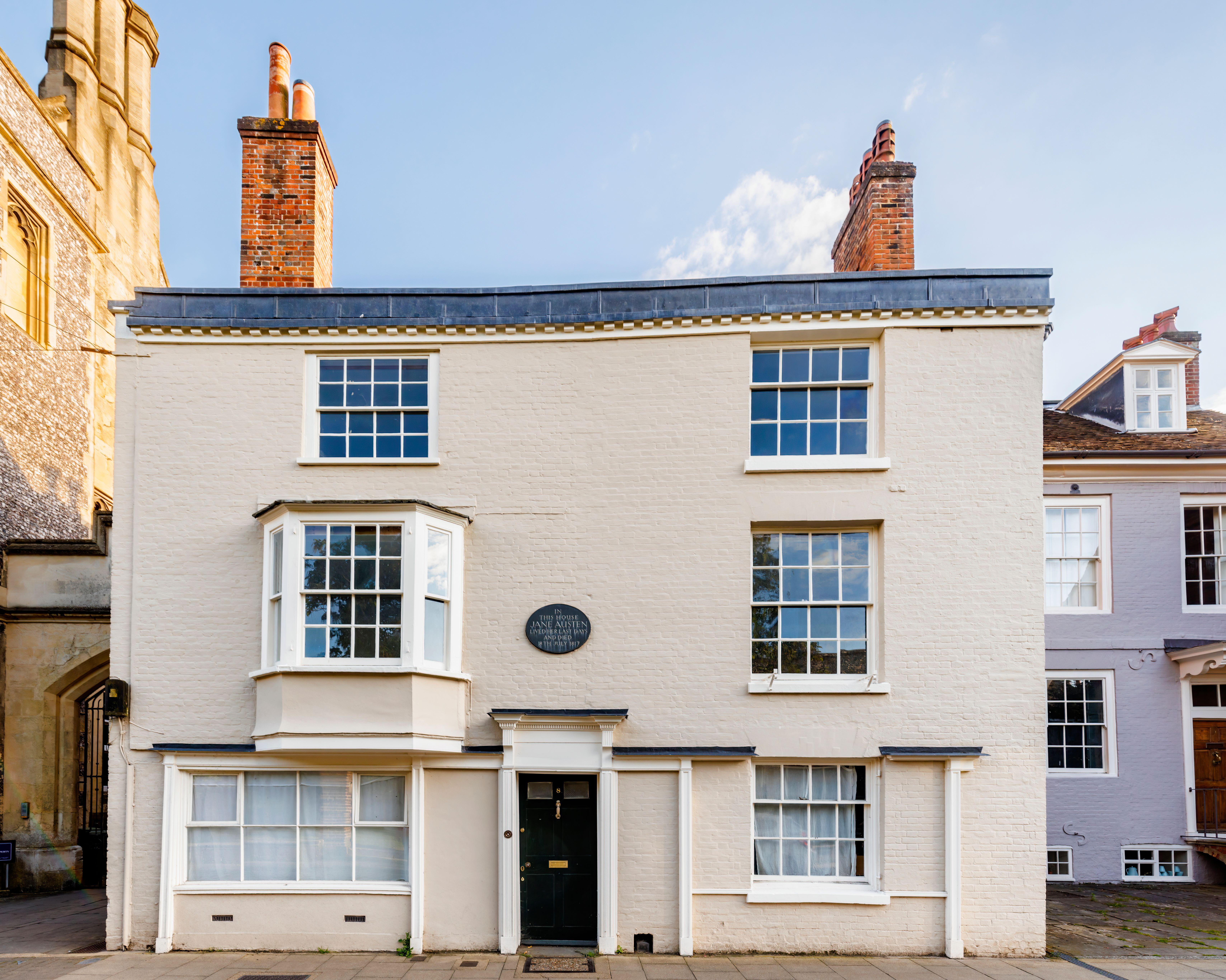 Front view of a beige brick two-story house with multiple white-framed windows, two tall red brick chimneys, and a black door with a plaque above it.