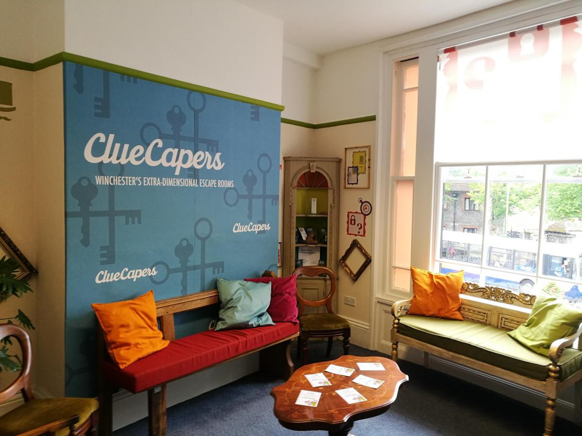 Cozy waiting area with wooden benches and chairs, colorful pillows, a coffee table with papers, and a blue wall featuring 'ClueCapers Winchester's Extra-Dimensional Escape Rooms' branding.