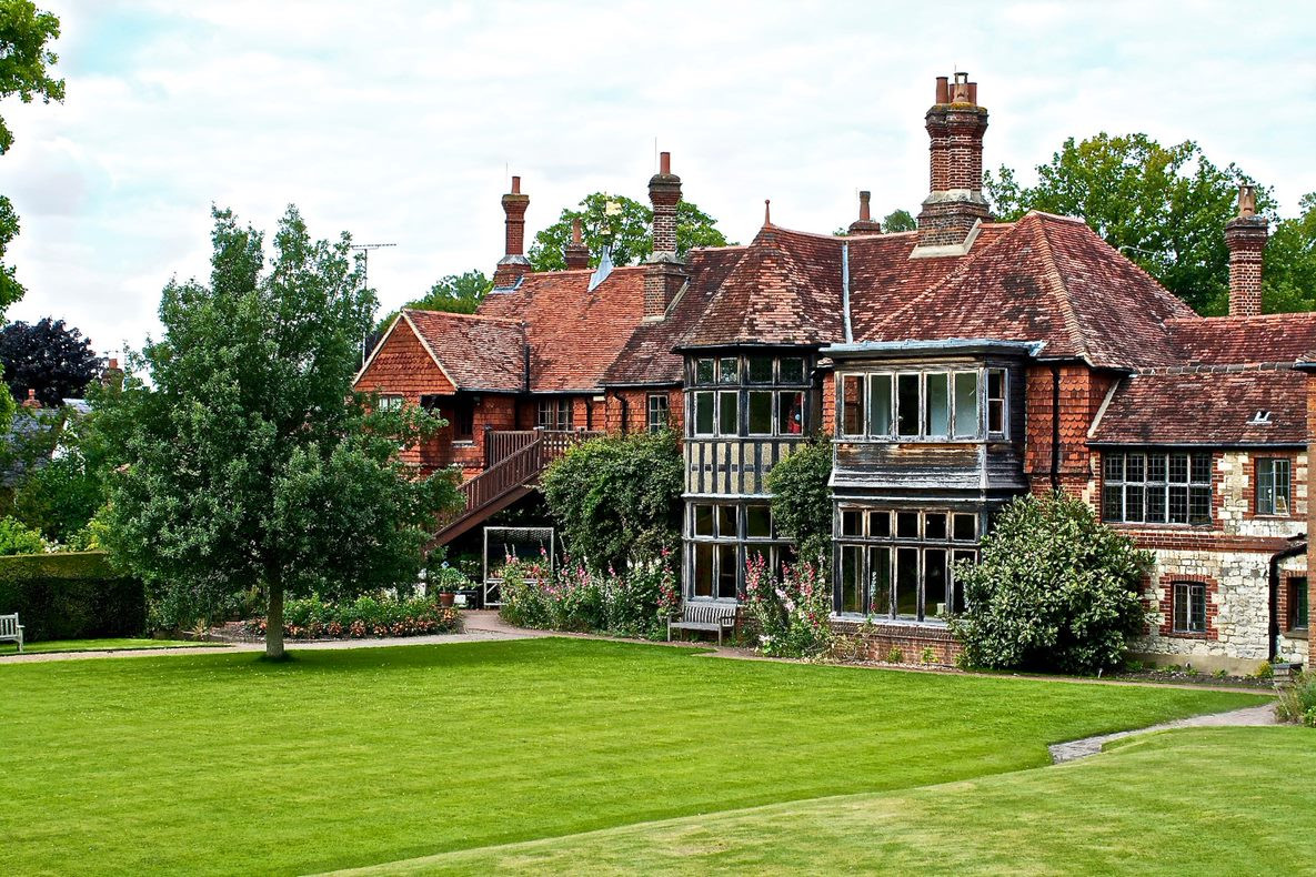 Large traditional English cottage with brick chimneys, wooden framed windows, surrounded by green lawn and trees.