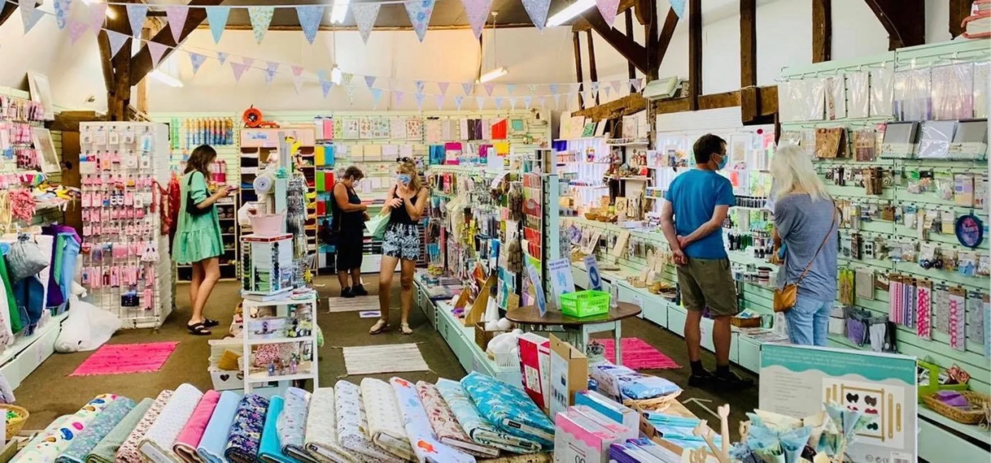 Interior of a craft store with fabric rolls displayed at the front and customers browsing various craft supplies on shelves and racks.