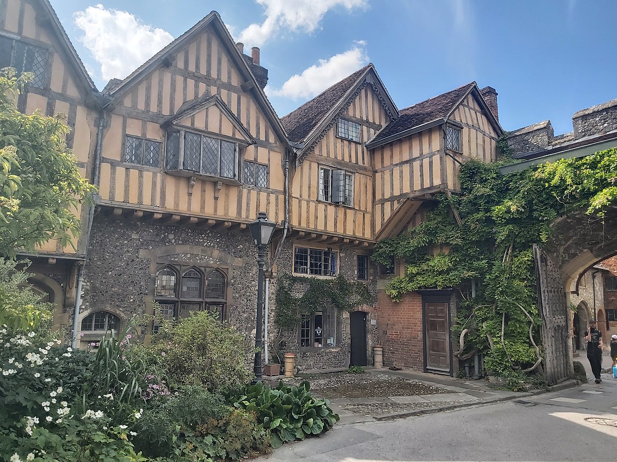 Historic timber-framed building with leaded glass windows, greenery, and an archway entrance under a blue sky.