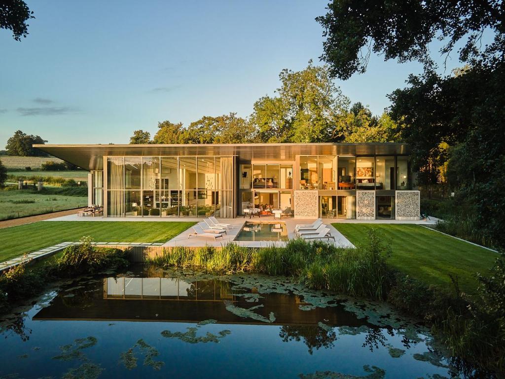 Modern glass and wood two-story house with lawn, pool, and pond in front, surrounded by trees.