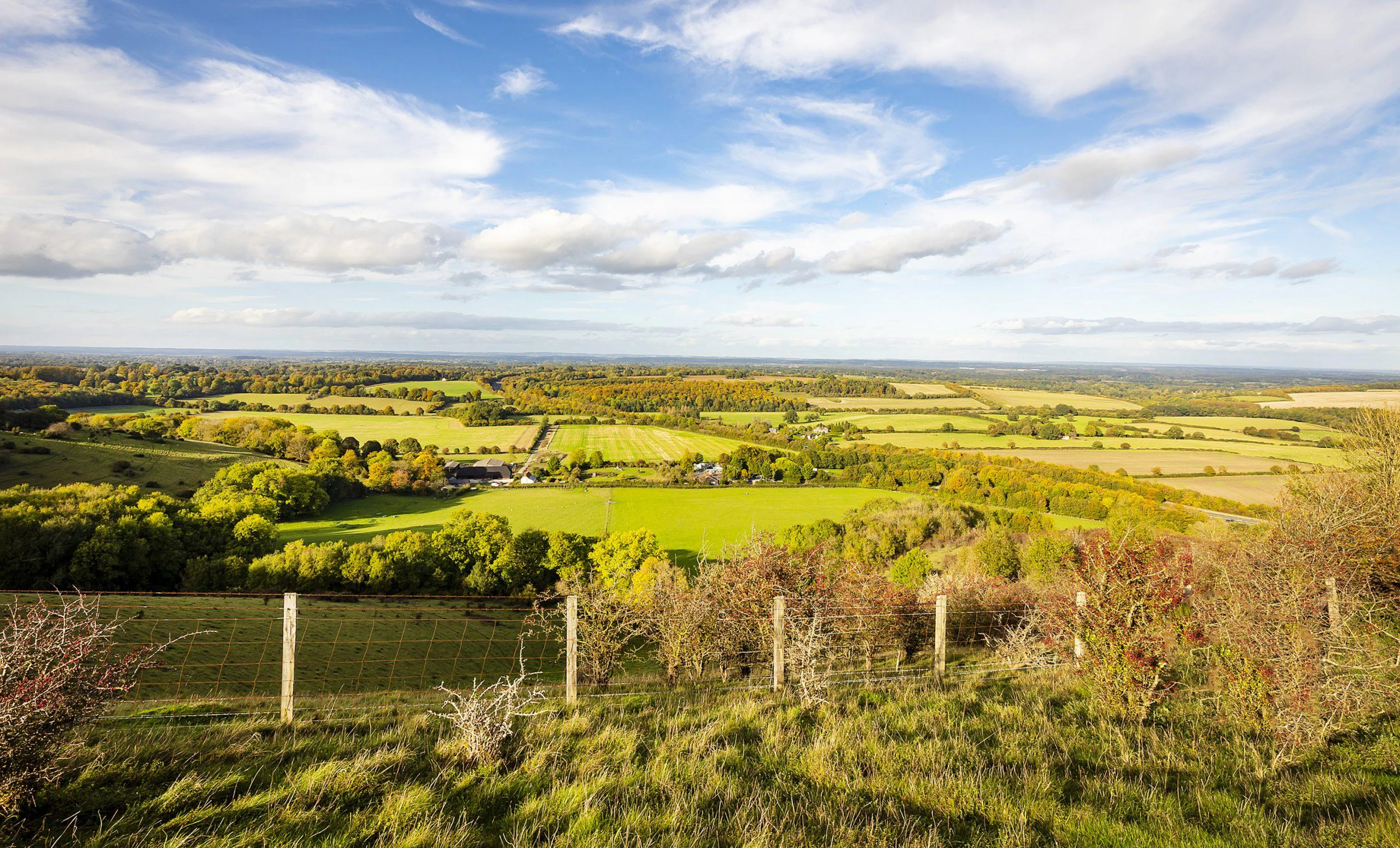 Scenic countryside view with green fields, trees, farm buildings, and a partly cloudy sky.