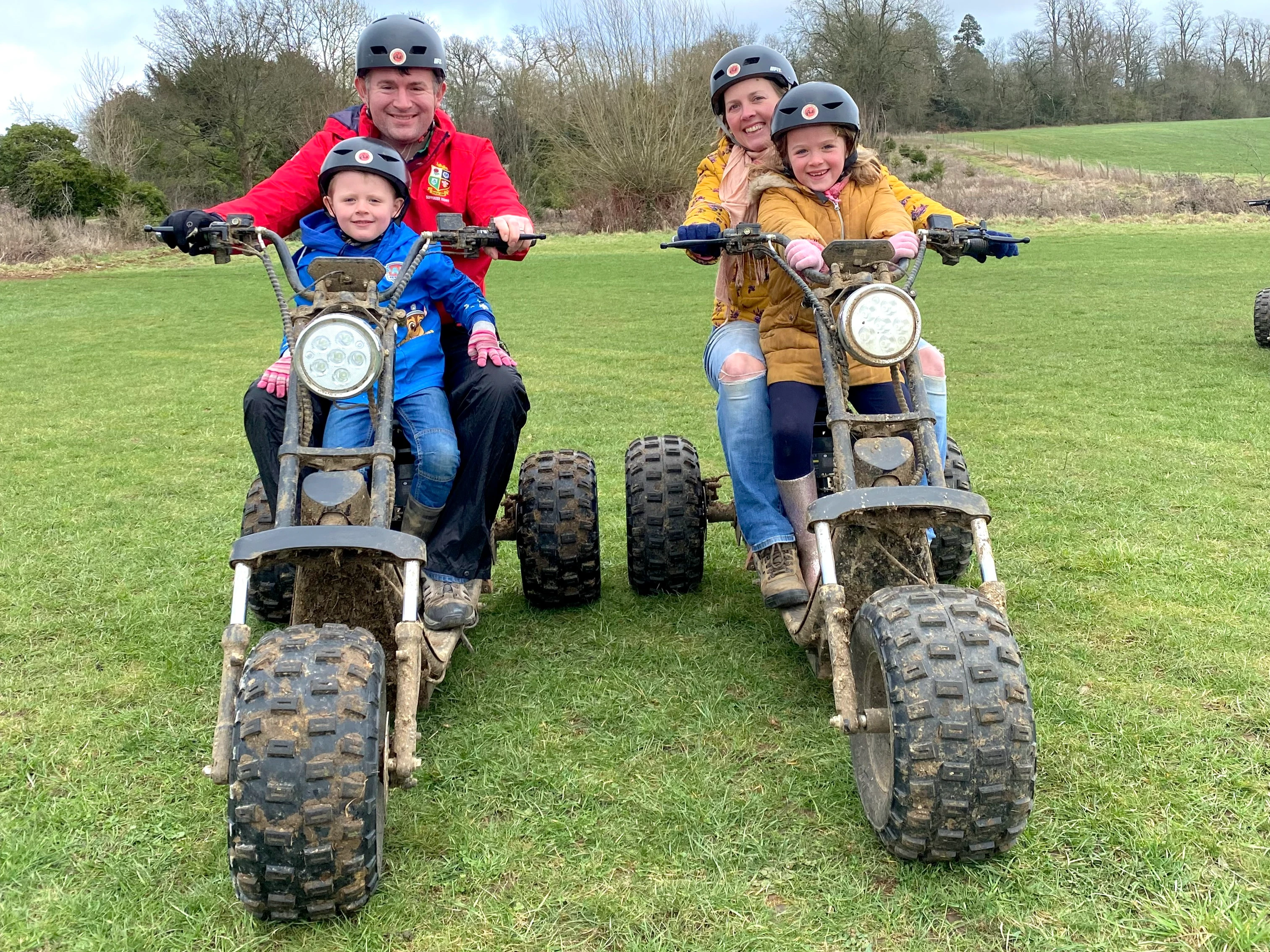 Two adults and two children wearing helmets sitting on muddy quad bikes on a grassy field.