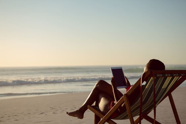 Person using a tablet on a beach chair by the sea representing travel, digital connection and lifestyle content Avant