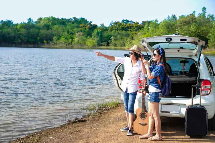 Two travelers stand beside an open rental car by the water with luggage, a guitar, and a camera, getting ready to explore more scenic stops with Avant