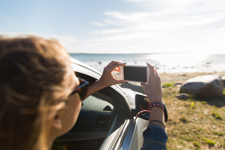 A traveler leans out from a parked car to photograph the shoreline and bright sea view, capturing the freedom of exploring coastal stops during a road trip with a rental car from Avant