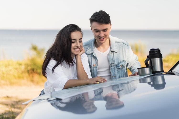 A couple stands by a car near the coast sharing a relaxed travel moment with drinks on the hood, reflecting the comfort of enjoying scenic stops and road trips with a rental car from Avant