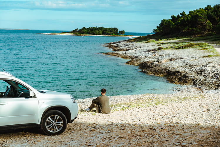 Un voyageur est assis à côté d’un SUV blanc sur un rivage rocheux face à une eau bleue paisible, reflétant la liberté de faire une pause, de se détendre et d’explorer des coins côtiers cachés avec une voiture de location Avant