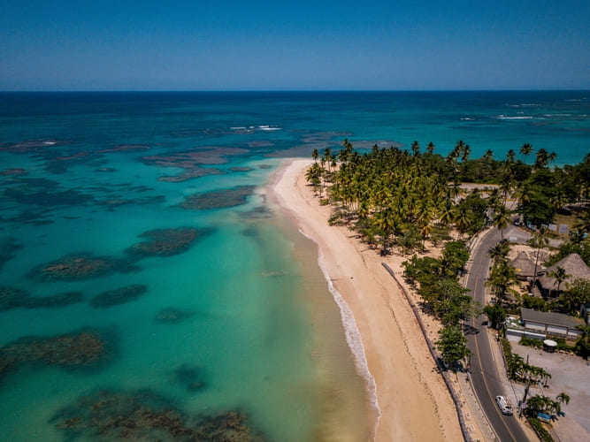 Une vue aérienne montre une plage tropicale avec une eau turquoise, des palmiers et une route côtière, invitant les voyageurs à découvrir des destinations en bord de mer à leur rythme avec la flexibilité d’Avant
