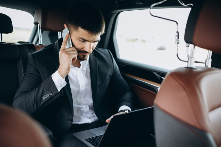 Un hombre de negocios trabaja en su laptop mientras atiende una llamada desde el asiento trasero de un vehículo, reflejando transporte ejecutivo que permite mantenerse productivo en movimiento con Avant