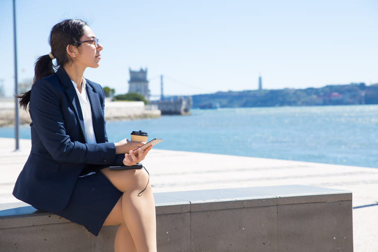 Una viajera de negocios está sentada frente al agua con un celular y café durante una pausa de trabajo, reflejando movilidad práctica para viajes corporativos y traslados en la ciudad con Avant