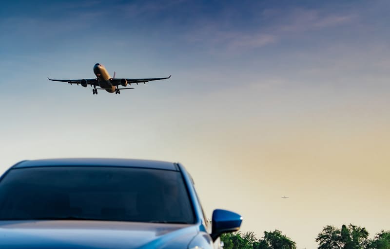 A car waits near an airport runway as an airplane flies overhead, highlighting a practical arrival or departure scene for travelers who need smooth airport mobility with Avant