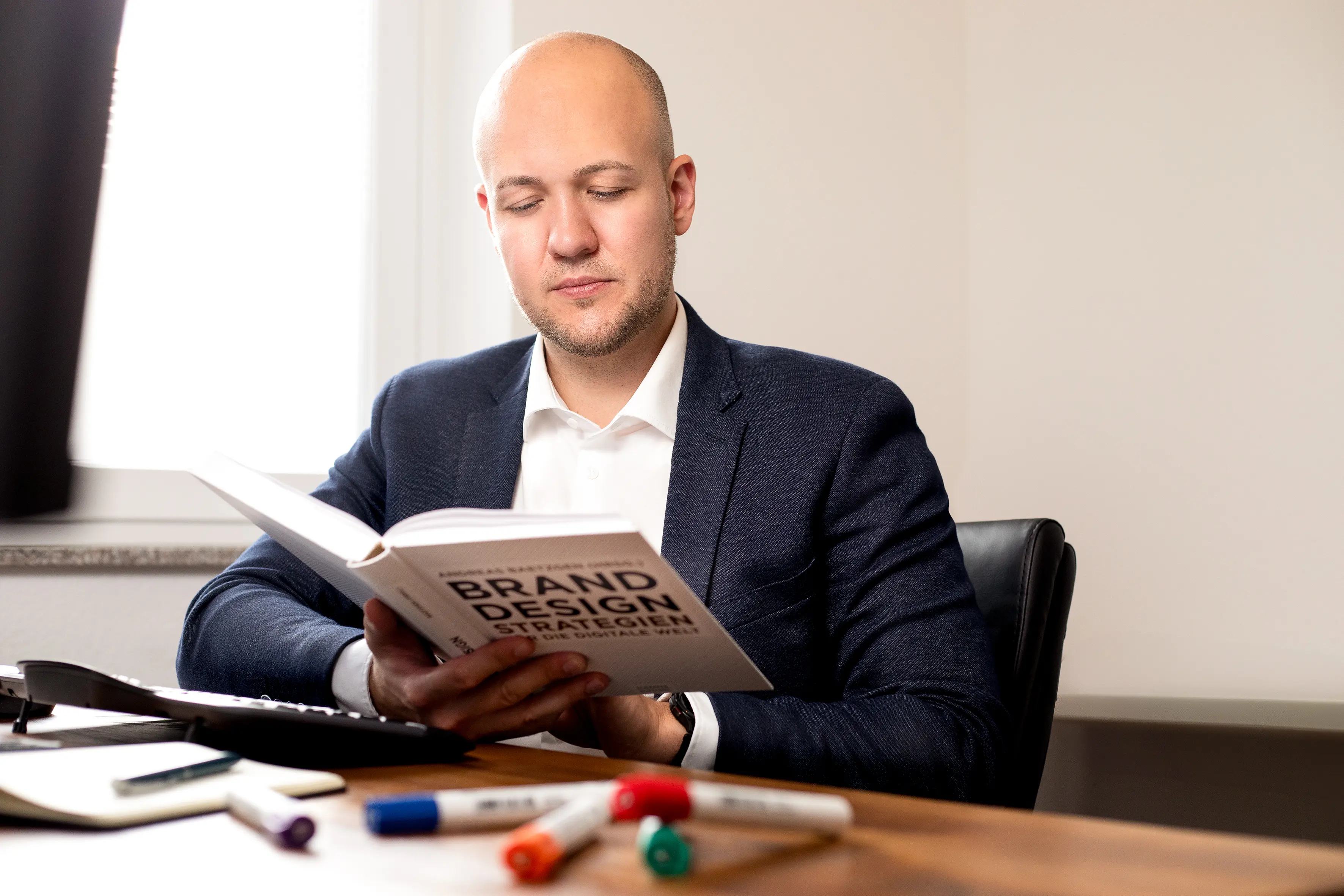 Matthias Hartmann, Gründer von Web Performance, in einem blauen Anzug und weißem Hemd sitzt an einem Tisch und hält ein Buch mit dem Titel "Brand Desing Strategien". Auf dem Tisch liegen mehrere Marker in verschiedenen Farben.