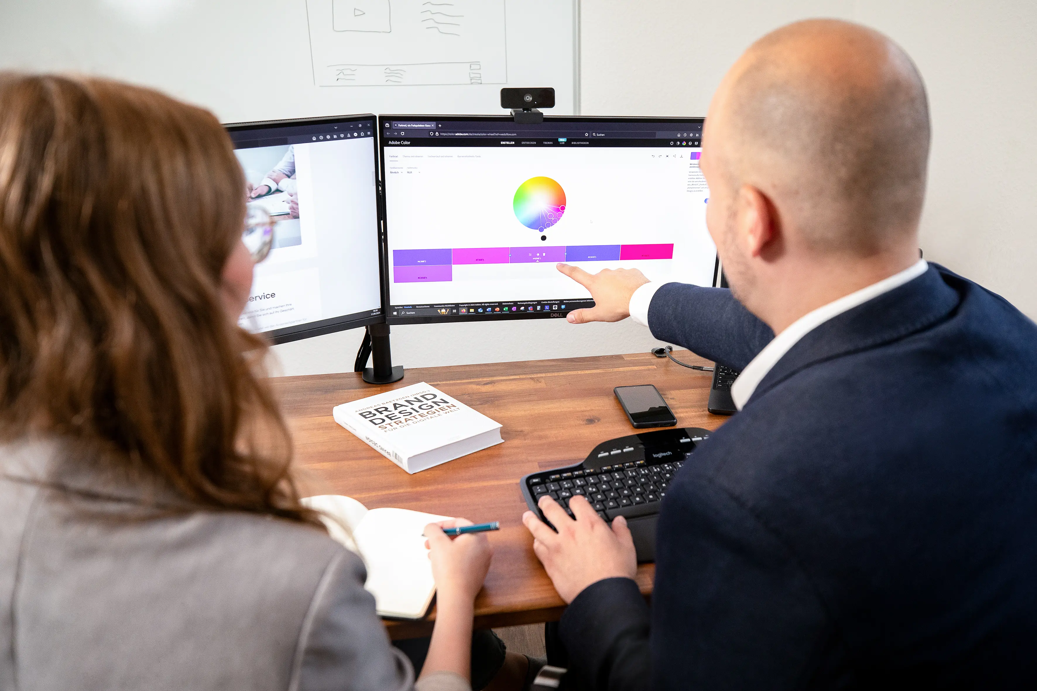 Matthias Hartmann, Gründer von Web Performance, im Anzug zeigt auf einen Bildschirm mit einer bunten Grafik. Eine Frau in einem grauen Blazer sitzt daneben und notiert etwas. Auf dem Tisch liegt ein Buch mit dem Titel "Brand Design Strategien"'.