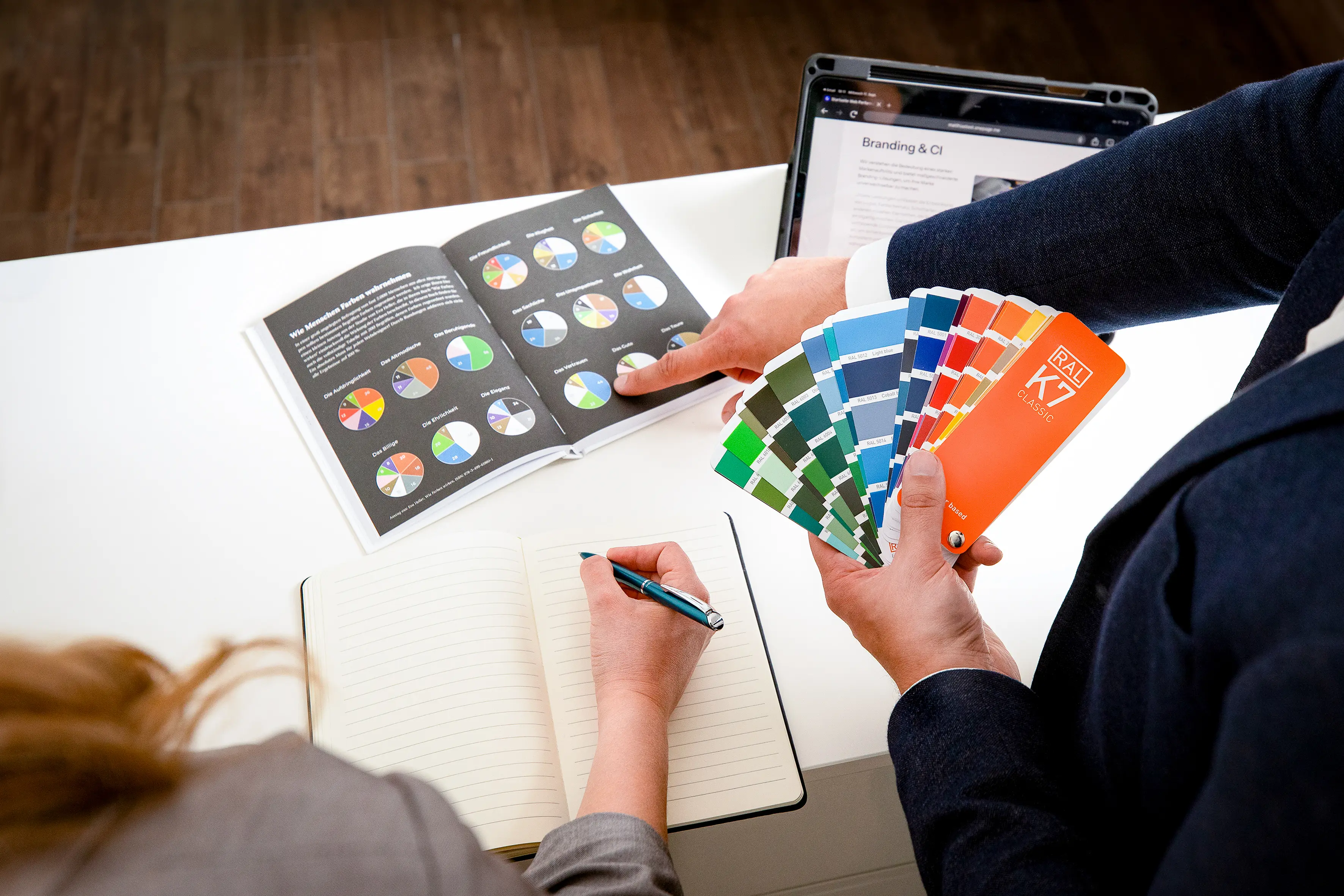 Matthias Hartmann, Gründer von Web Performance, zeigt RAL K7 Farbpaletten in der Hand und weist auf ein Diagramm in einem Branddesign-Buch. Eine Mitarbeiterin schreibt in ein Notizbuch.