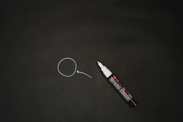 Ein schwarzer Hintergrund mit einem weißen Kreis und einem Pfeil, der auf den Kreis zeigt. Daneben liegt ein schwarzer Marker mit weißer Spitze.