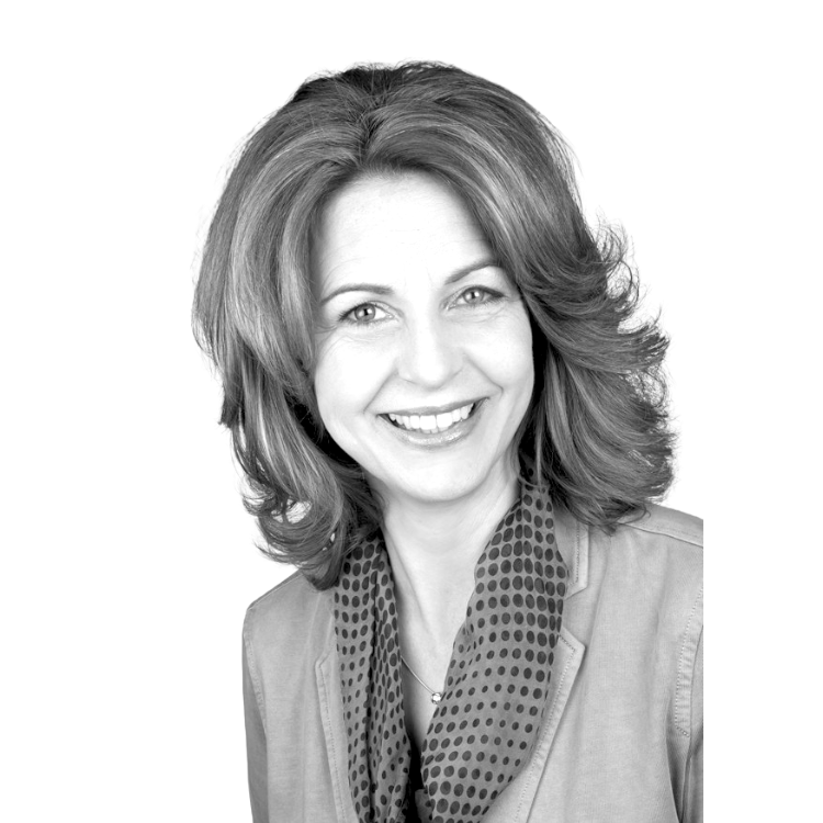 Margit Bloch von Praxis Margit Bloch mit schulterlangem, gewelltem Haar, trägt gepunkteten Schal und hellen Blazer, vor schwarzem Hintergrund.