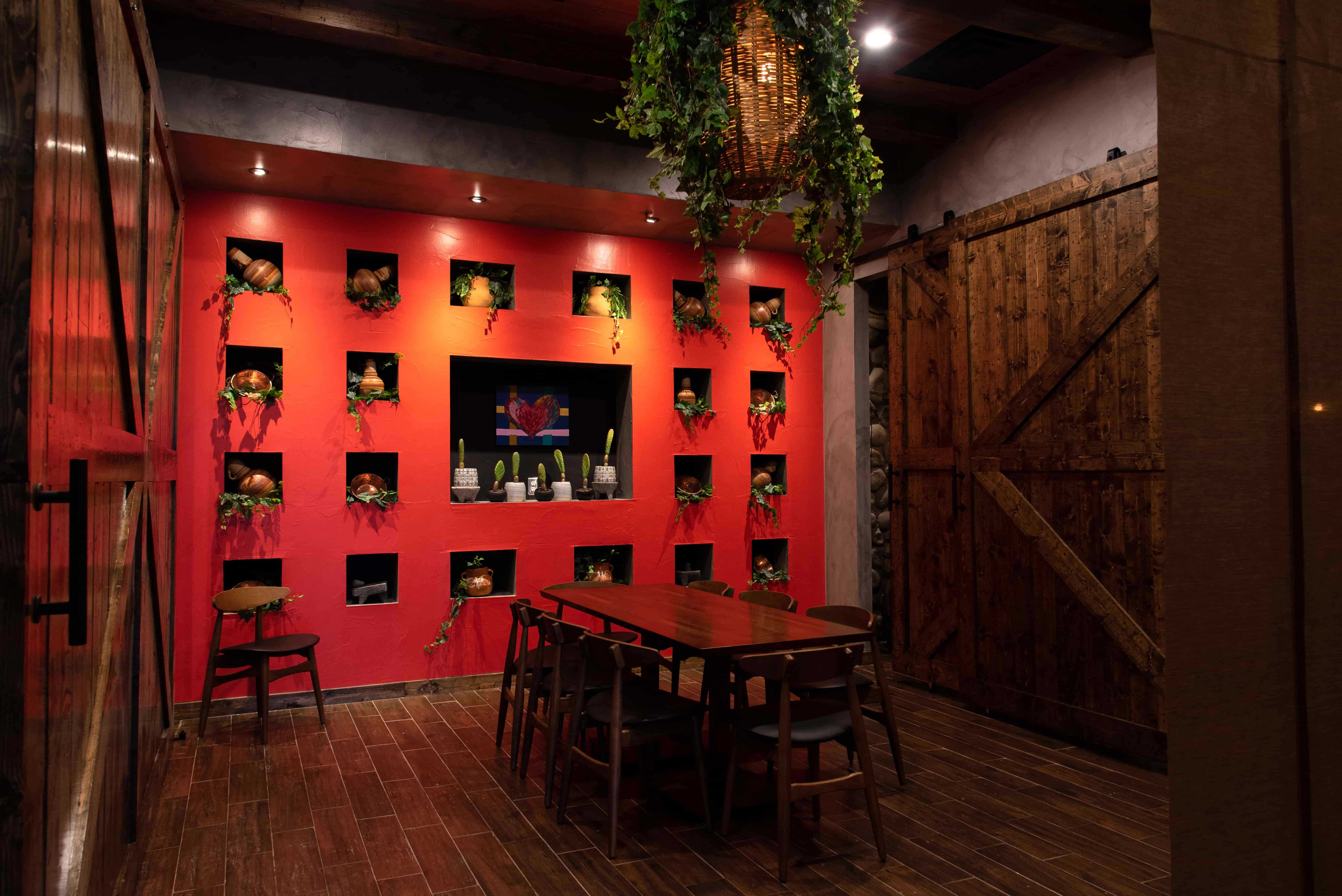 Dining room with red wall featuring pottery and plants in square niches and wooden table with six chairs.