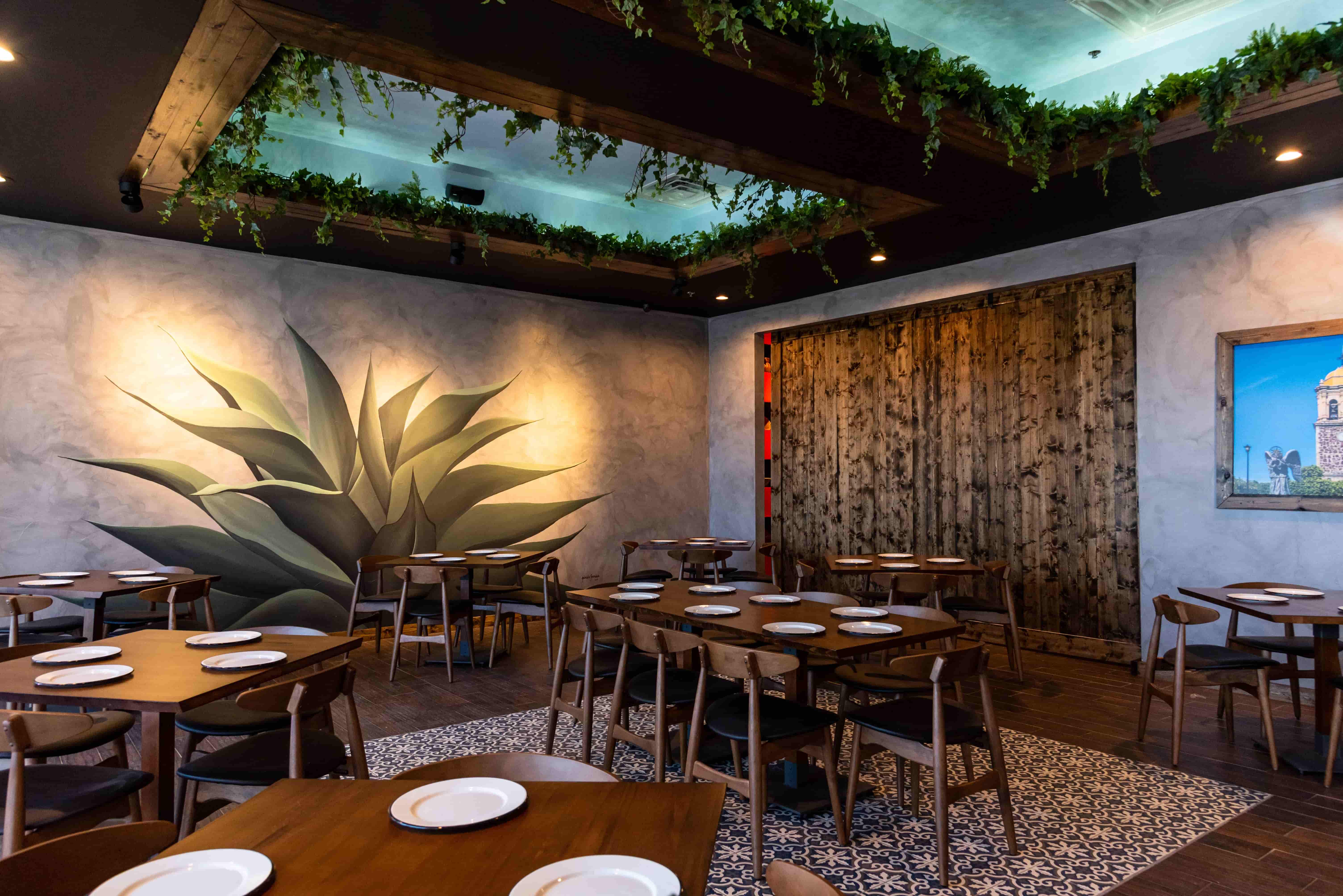 Restaurant interior with wooden tables, chairs, white plates, plant mural, and greenery on ceiling beams.
