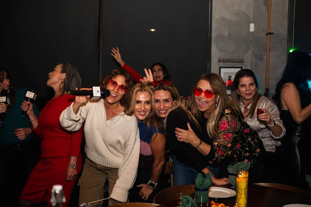 Group of women smiling and posing indoors at a social gathering with drinks and microphones.