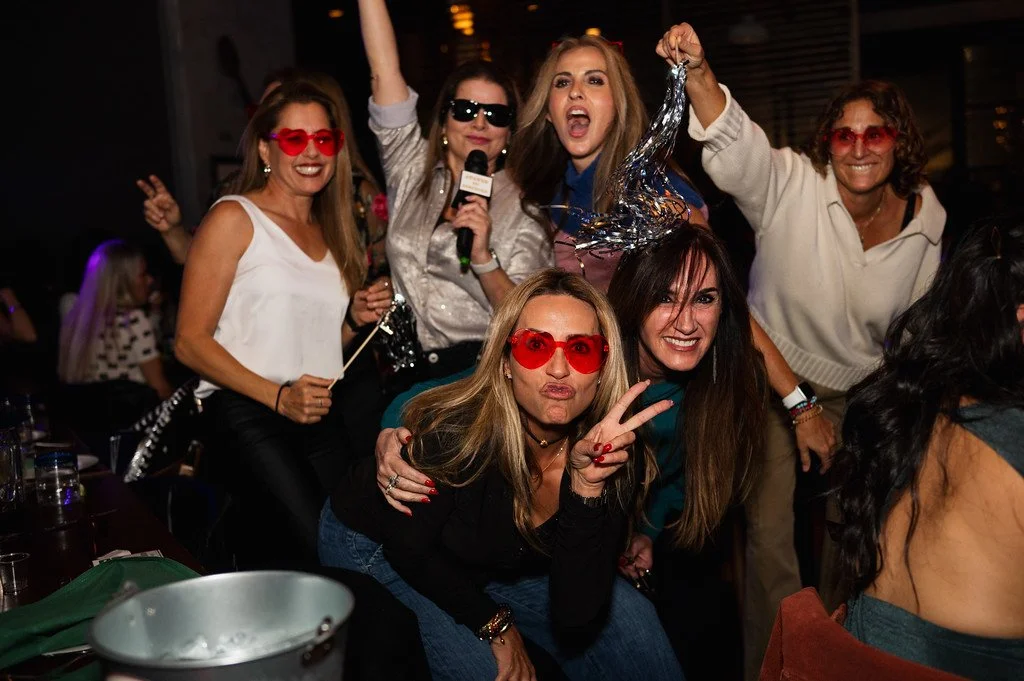 Group of six women posing energetically at a nighttime party, some wearing red heart-shaped glasses.