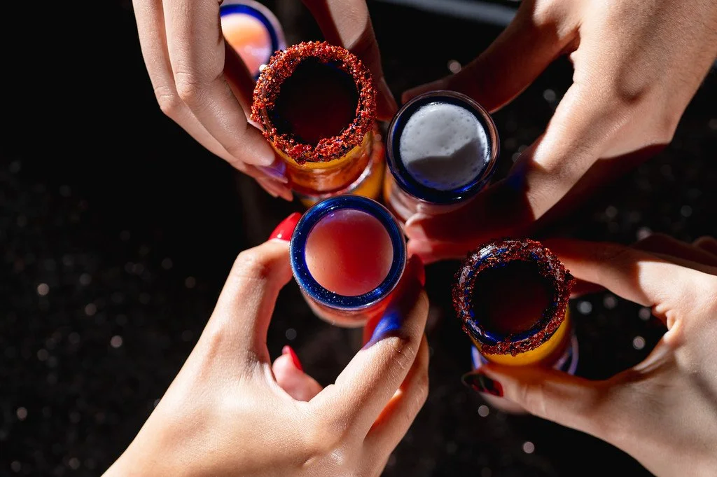 Four hands holding and toasting bottles with chamoy rims, photographed from above on black background.