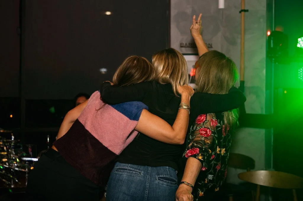 Three women hugging each other at a dimly lit indoor party with green lighting.
