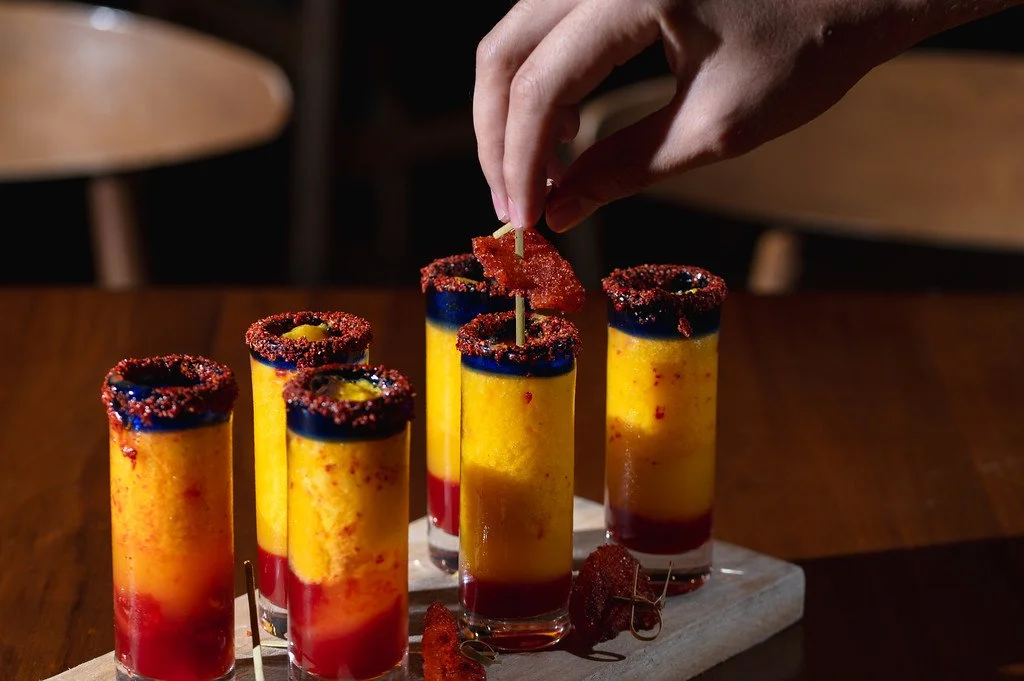 Hand placing a chili-coated garnish on a yellow and red layered drink with chili-rimmed glasses.