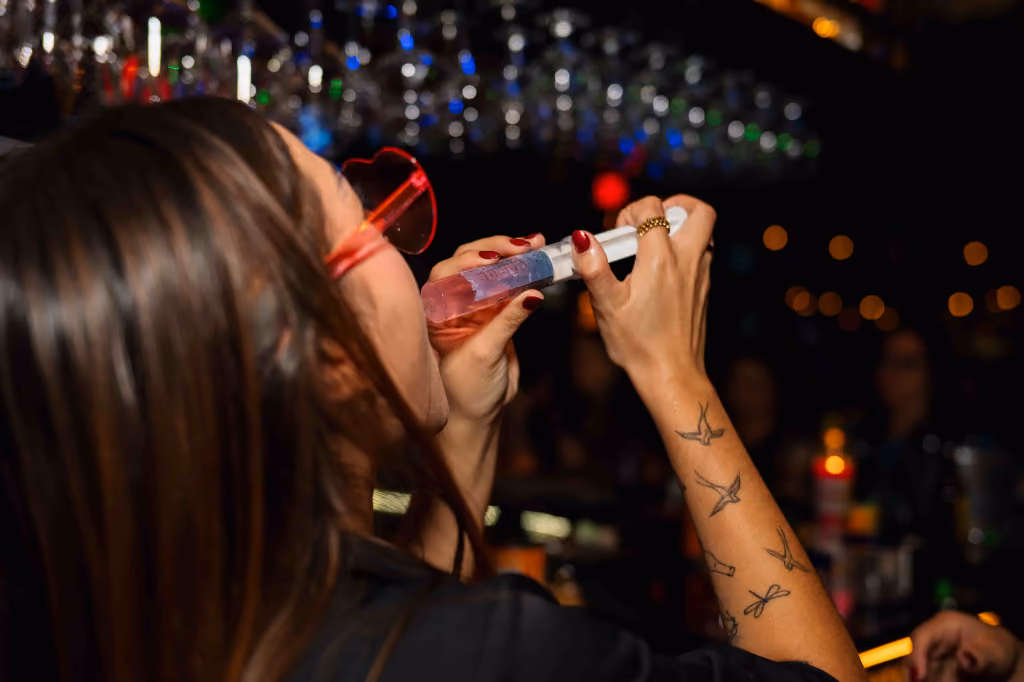 Woman with tattoos and red sunglasses drinks from a syringe at a dimly lit bar.