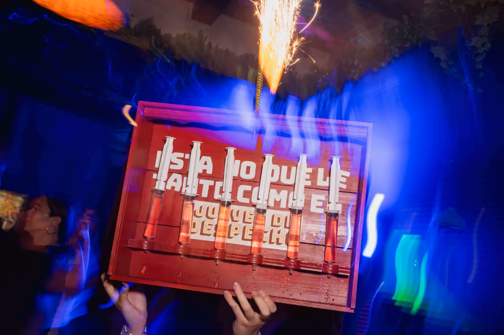 Hands hold a red tray with six syringes filled with orange liquid under a sparkler at a party.