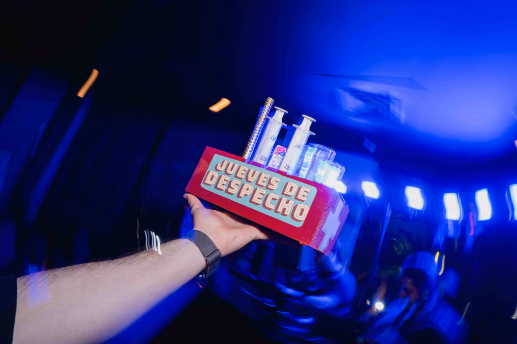 Arm holding a tray with syringes and glasses labeled 'Jueves de Despecho' under blue lighting.