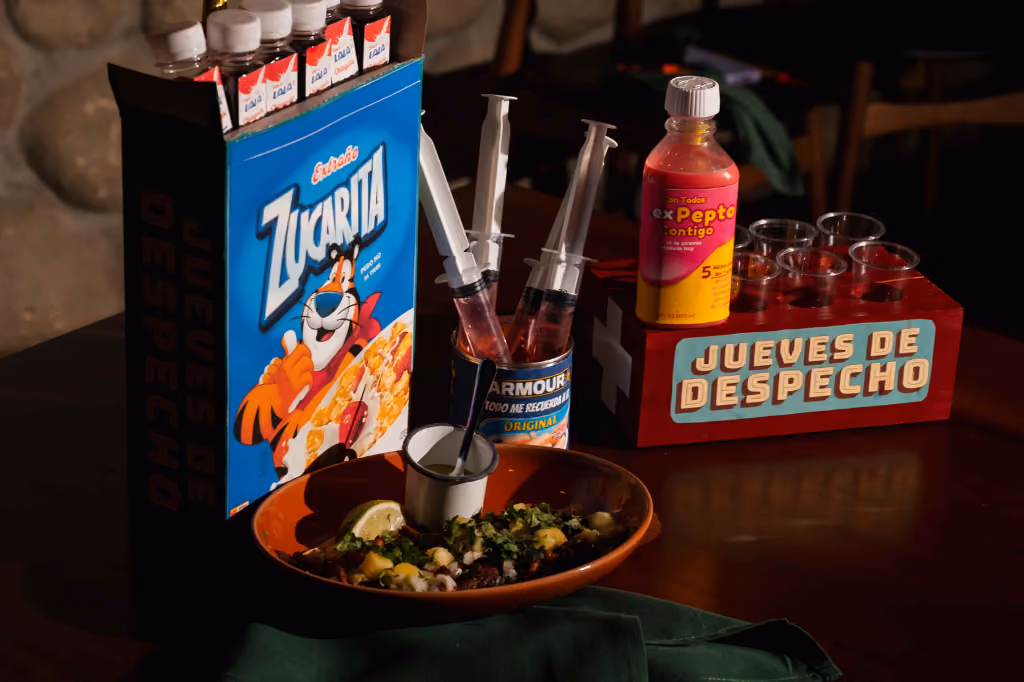 Plate of food with lime, syringes filled with red liquid, a cereal box, and a Pepto bottle on a table.
