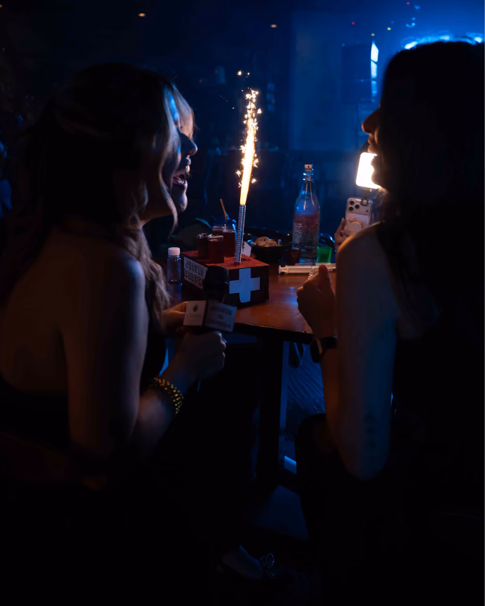 Two women smiling and sitting at a table with a lit sparkler in a dark setting.