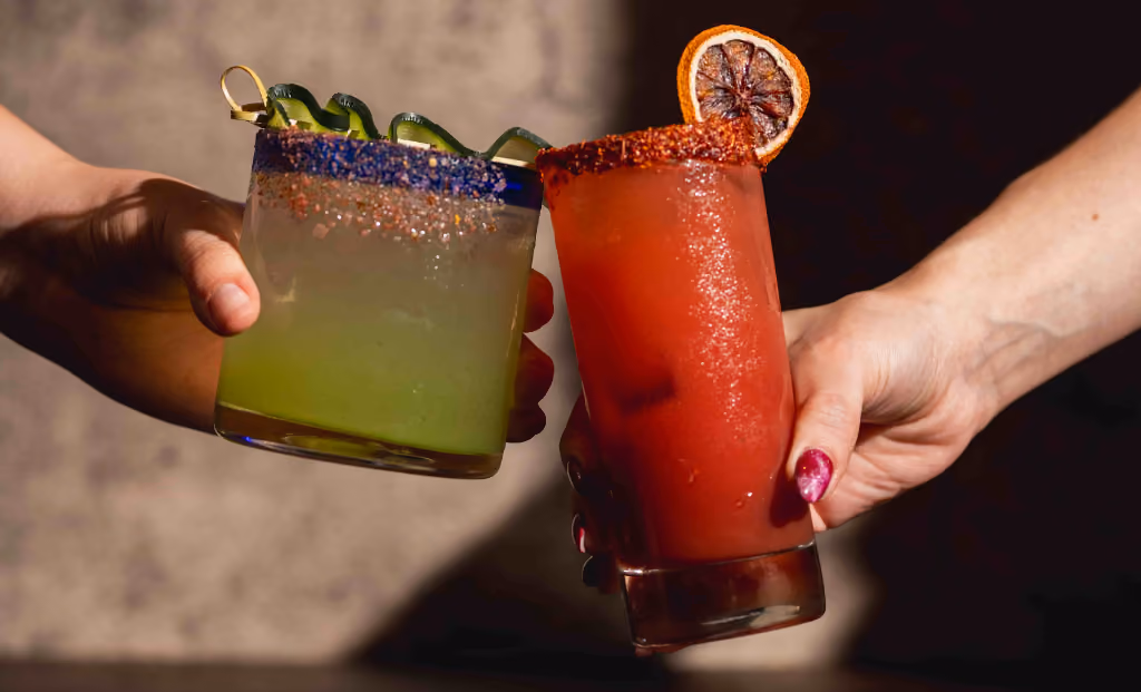 Two hands holding cocktails: one green with cucumber garnish, one red with dried citrus garnish.
