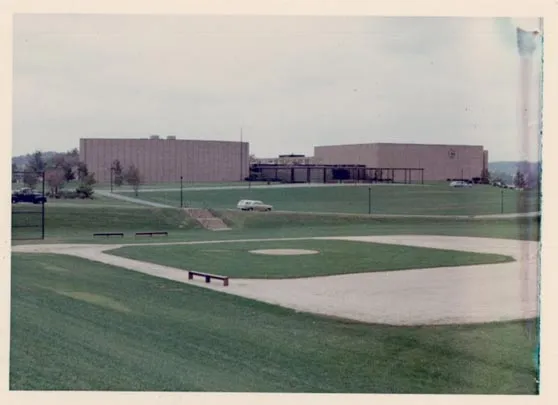 View of a large school building with a baseball field in the foreground under a cloudy sky.