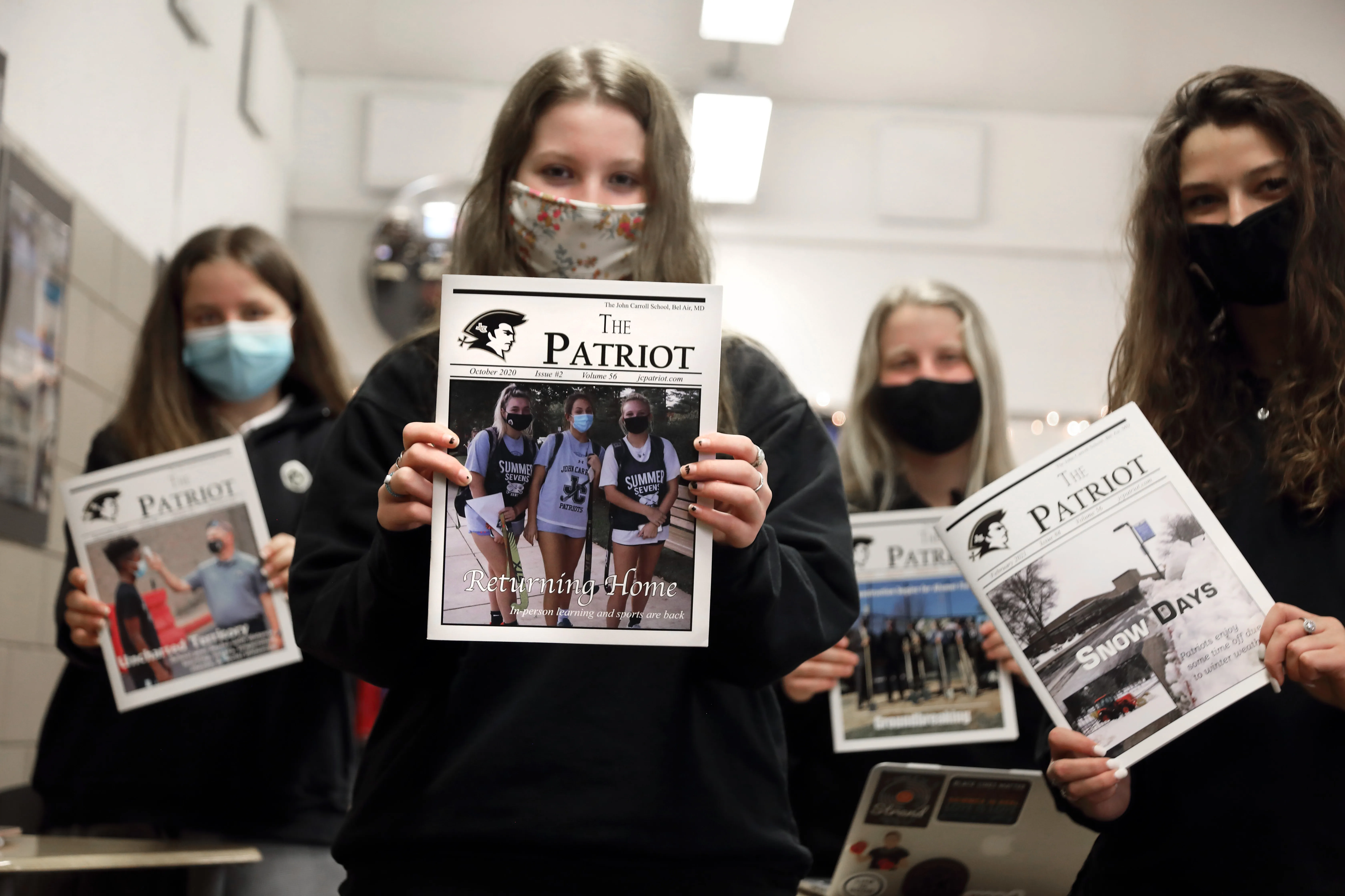 Four masked students holding up issues of The Patriot school newspaper inside a hallway.