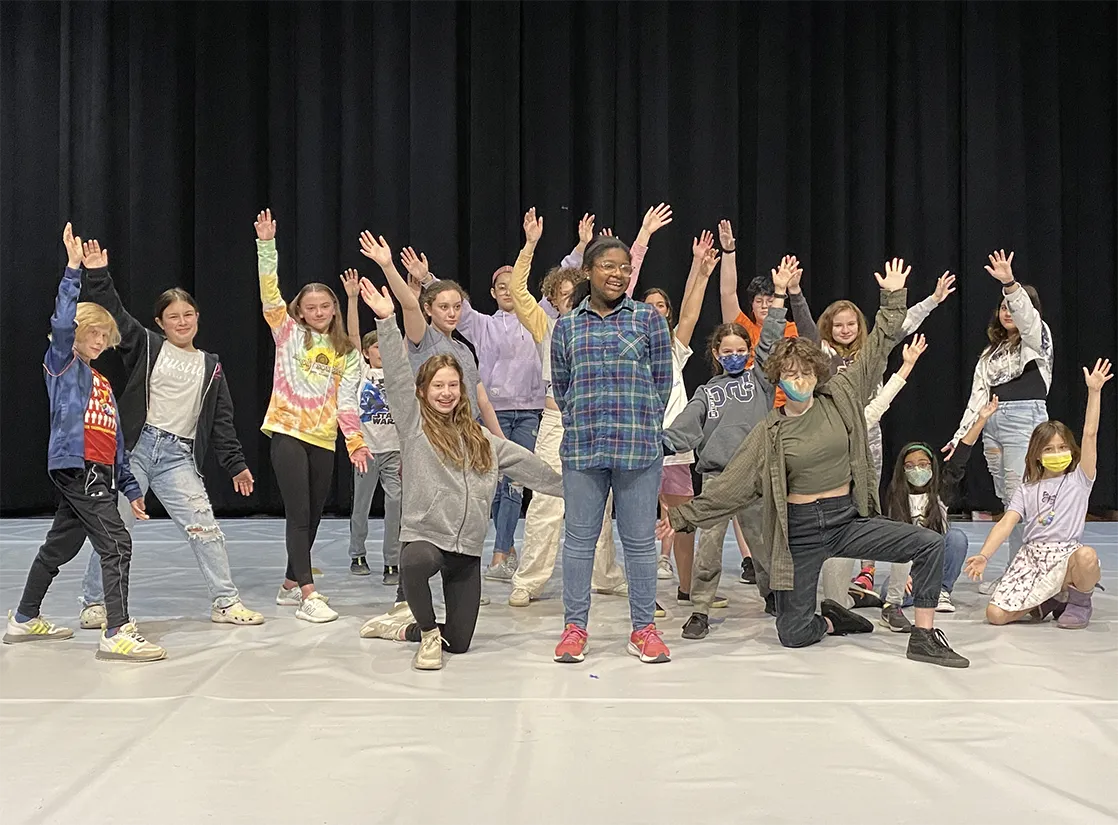 Group of children and teenagers smiling and posing with raised hands on a stage with a black curtain backdrop.