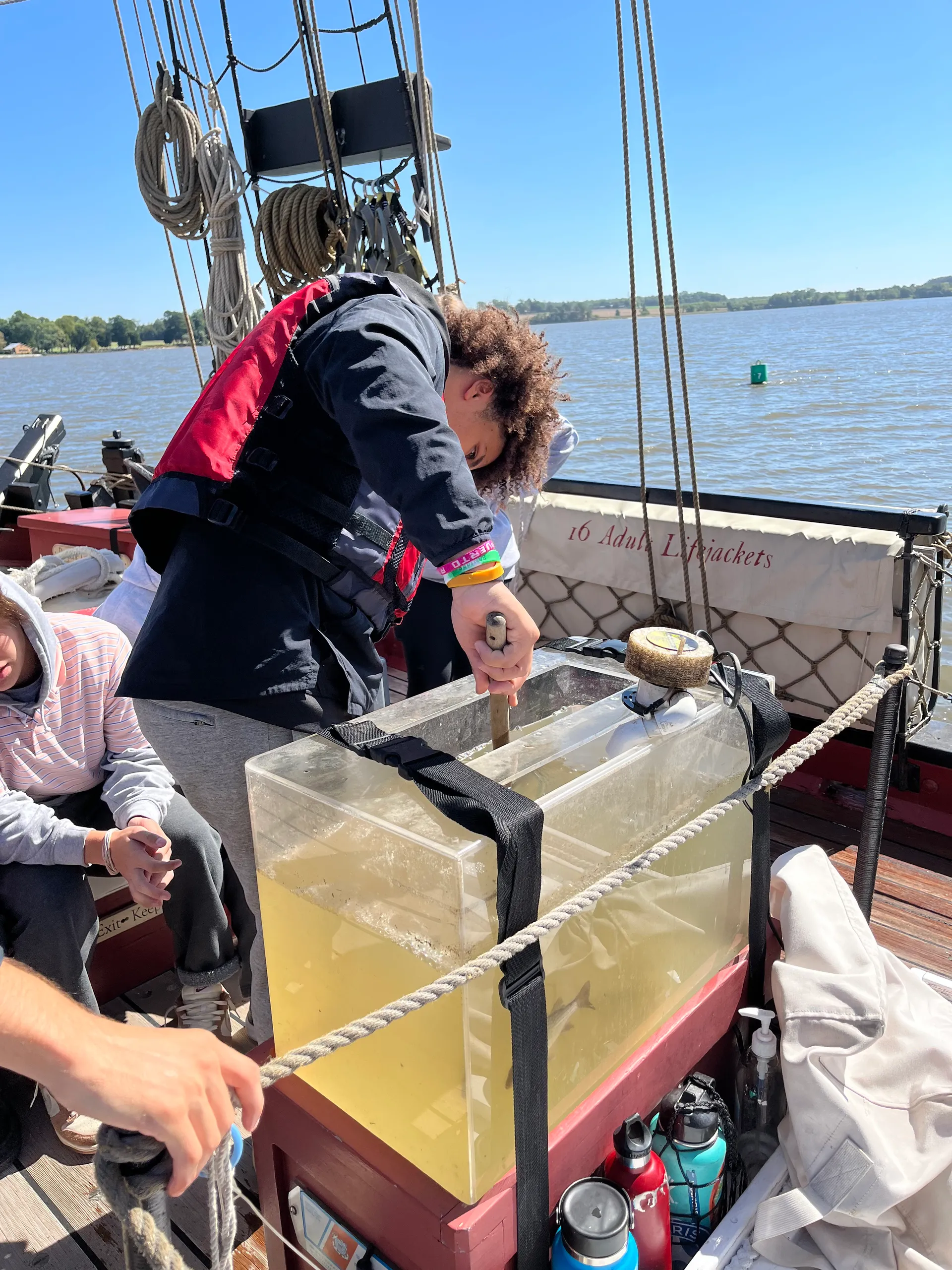Young person in a life jacket leaning over a large clear tank filled with water on a boat deck, stirring the water with a wooden stick.