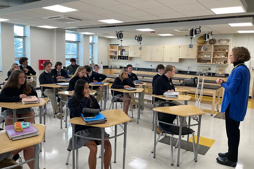 A classroom with students seated at individual desks listening to a standing teacher wearing a blue cardigan.