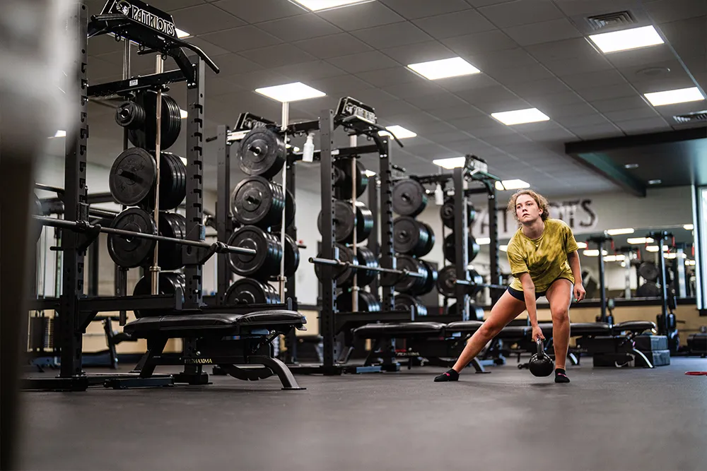 Young woman in yellow shirt performing side lunge with kettlebell in a gym filled with weightlifting equipment.