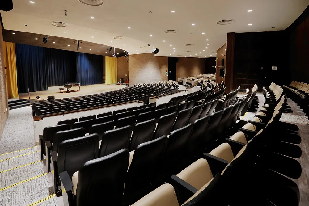 Empty auditorium with rows of black and beige seats facing a stage with a grand piano and closed blue and yellow curtains.