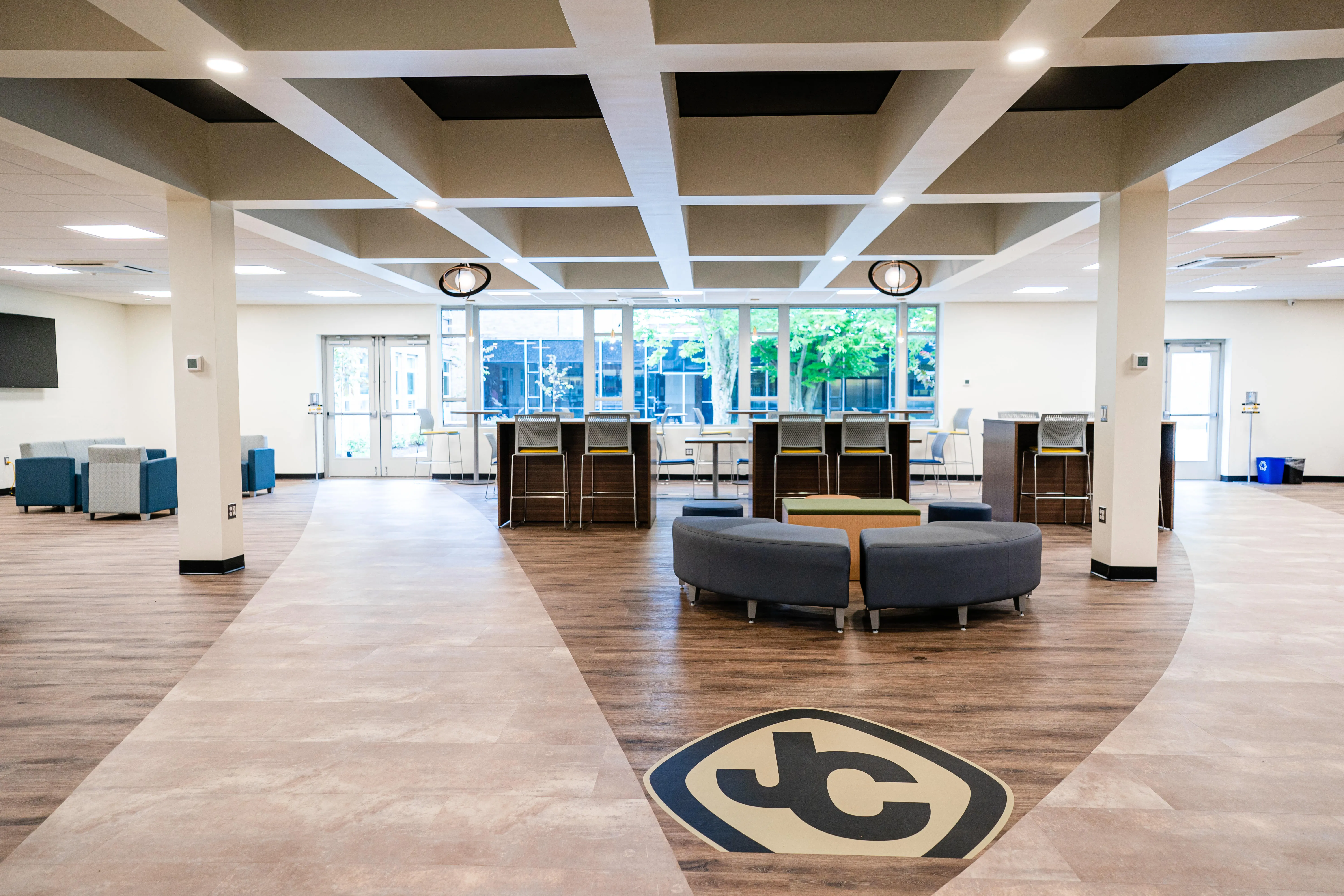 Modern open lounge area with curved seating, tall tables with chairs, large windows, and a JC logo on the wooden floor.