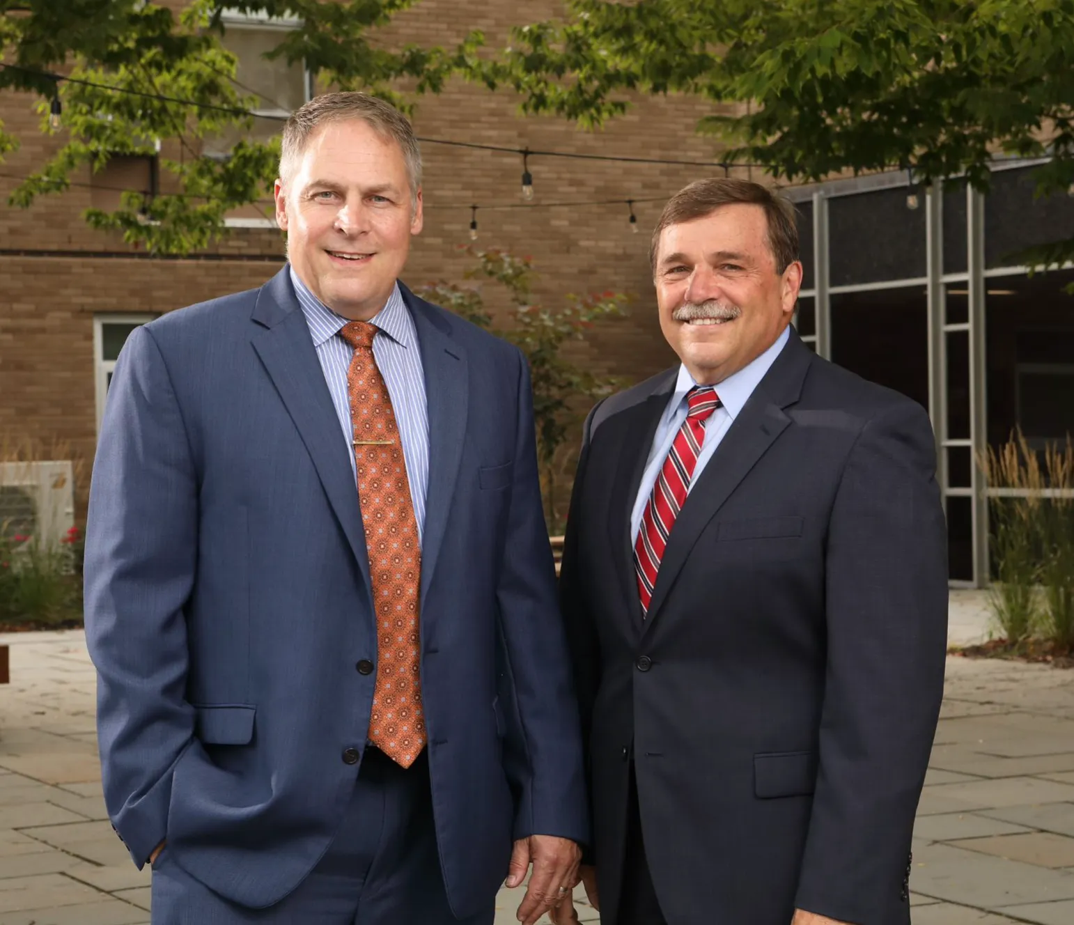 Two middle-aged men in business suits smiling outdoors with trees and a brick building in the background.