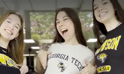 Three smiling young women wearing college-themed shirts, with the center woman showing her Georgia Tech shirt.