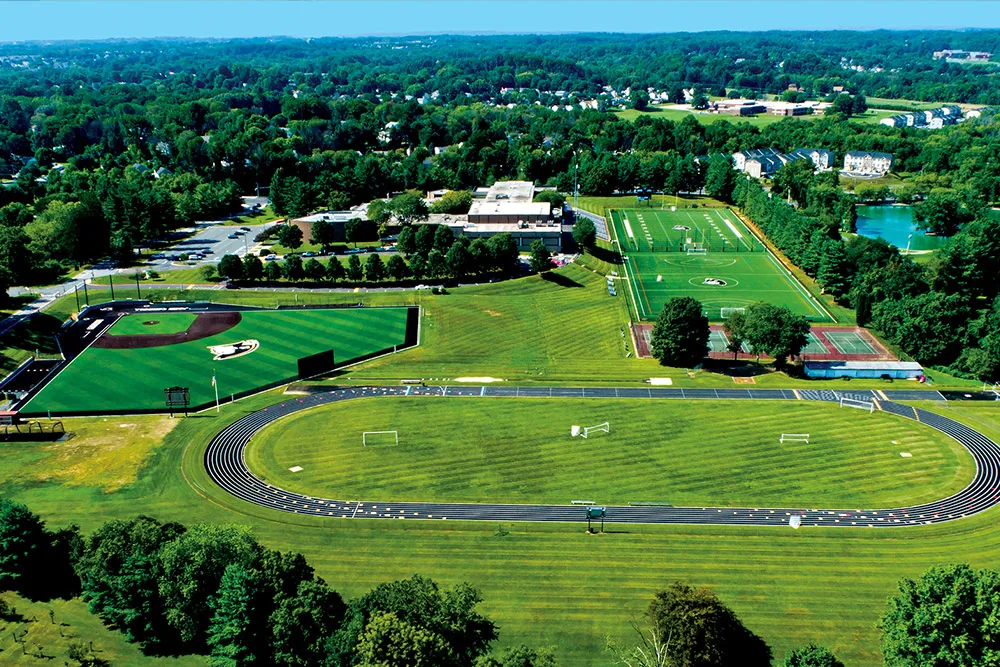 Aerial view of a sports complex featuring a running track surrounding a grass field, a baseball diamond, a soccer field, tennis courts, and surrounding trees.