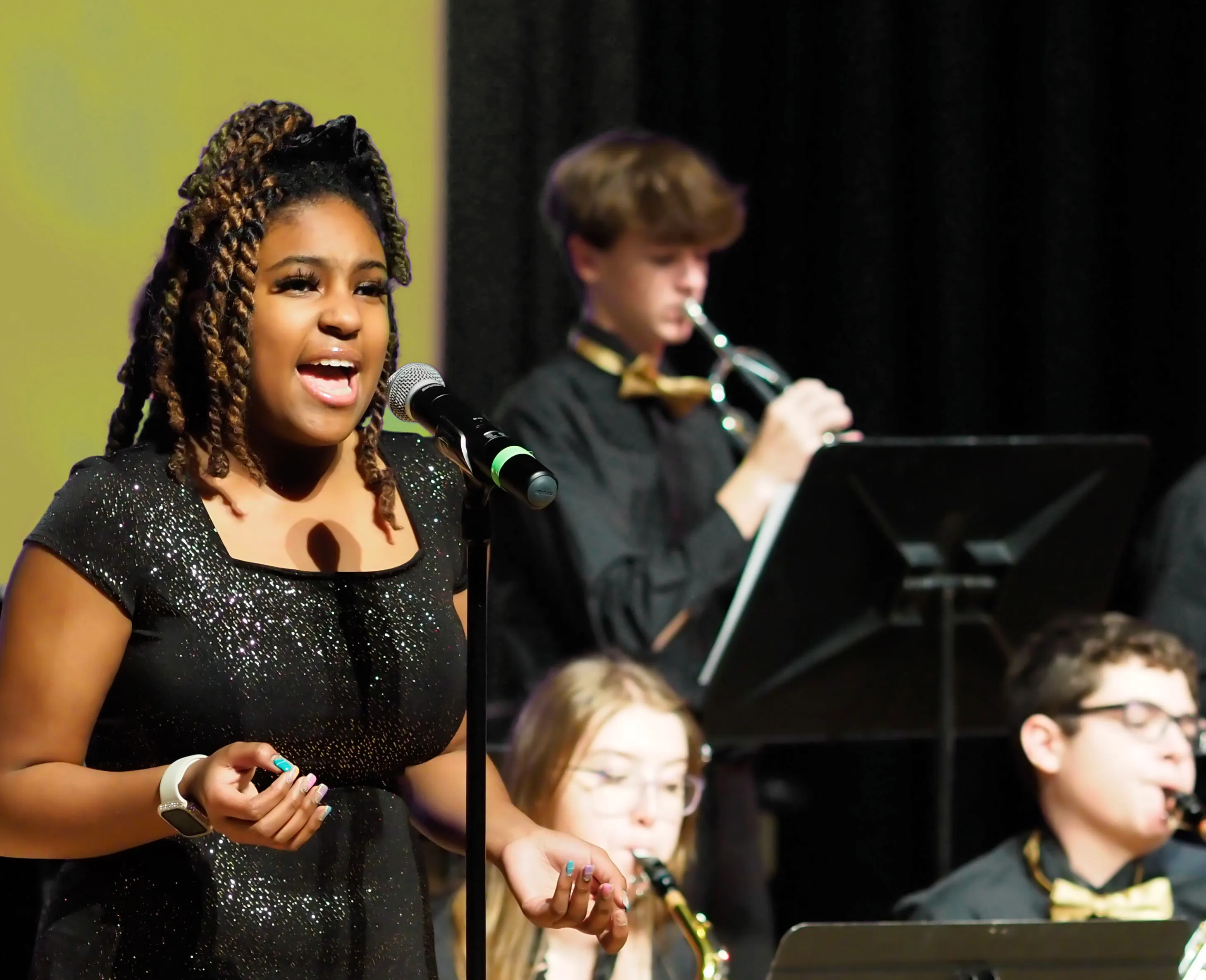 A young woman singing into a microphone with three musicians playing brass and woodwind instruments in the background.