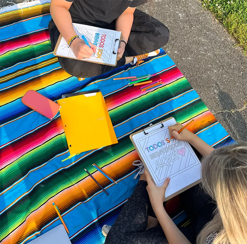 Two people sitting on a colorful striped blanket outdoors, coloring worksheets that say 'TODOS SOY BIENVENIDOS AQUÍ' with colored pencils scattered around.