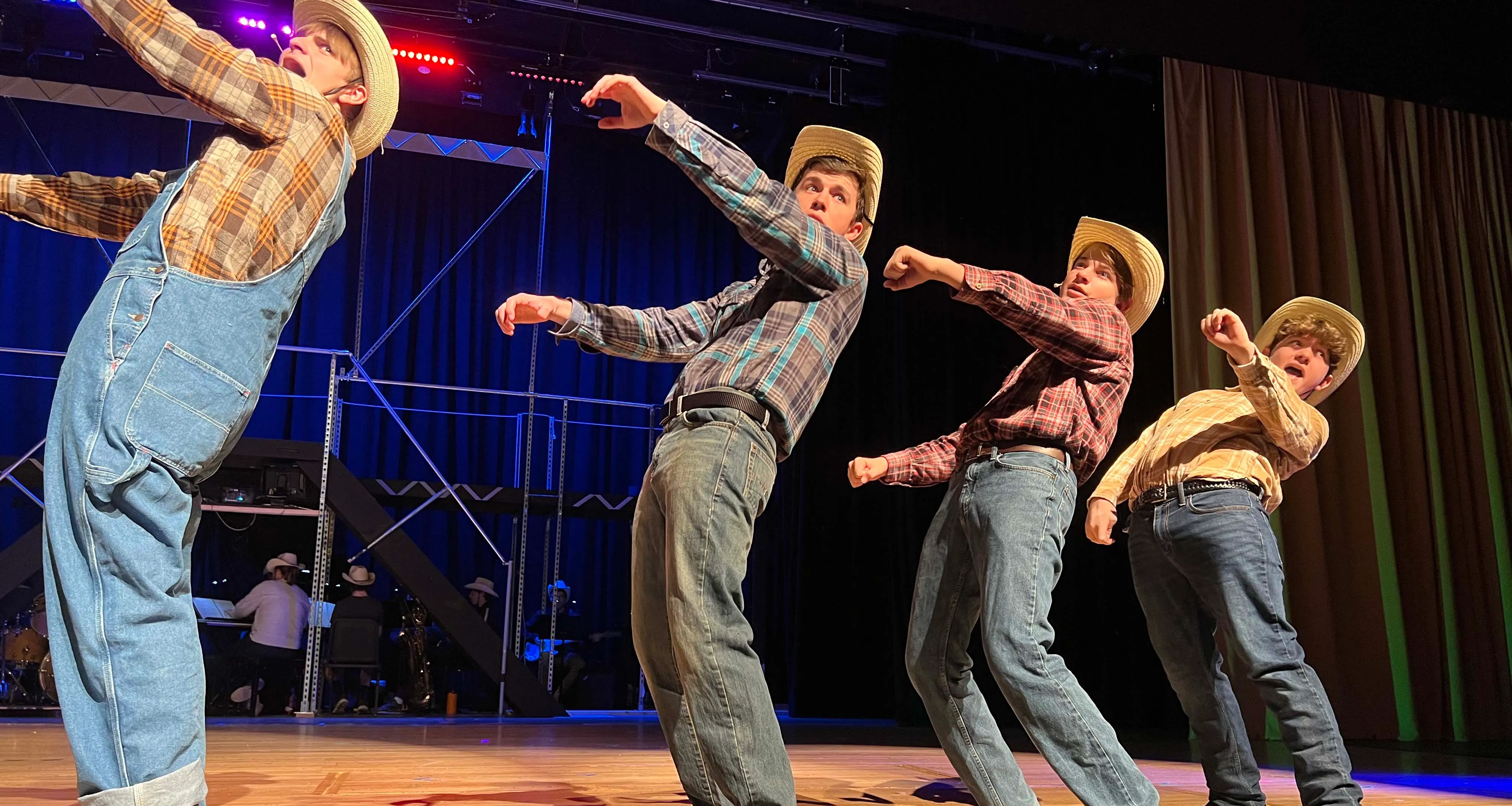 Four young men in plaid shirts, jeans, and cowboy hats performing a dance on stage with dramatic leaning poses.
