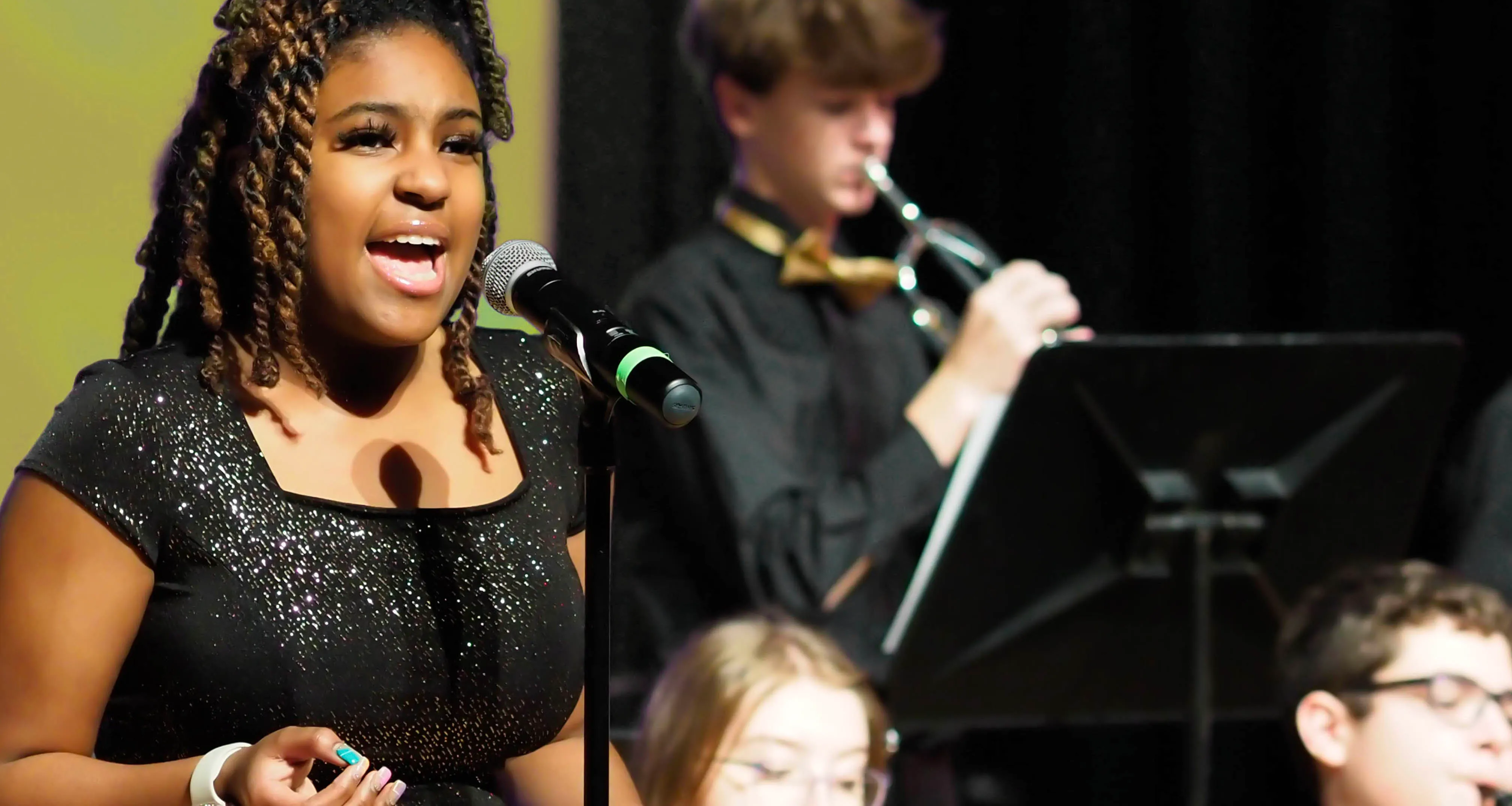 Young woman singing into a microphone with a trumpet player and other musicians blurred in the background.